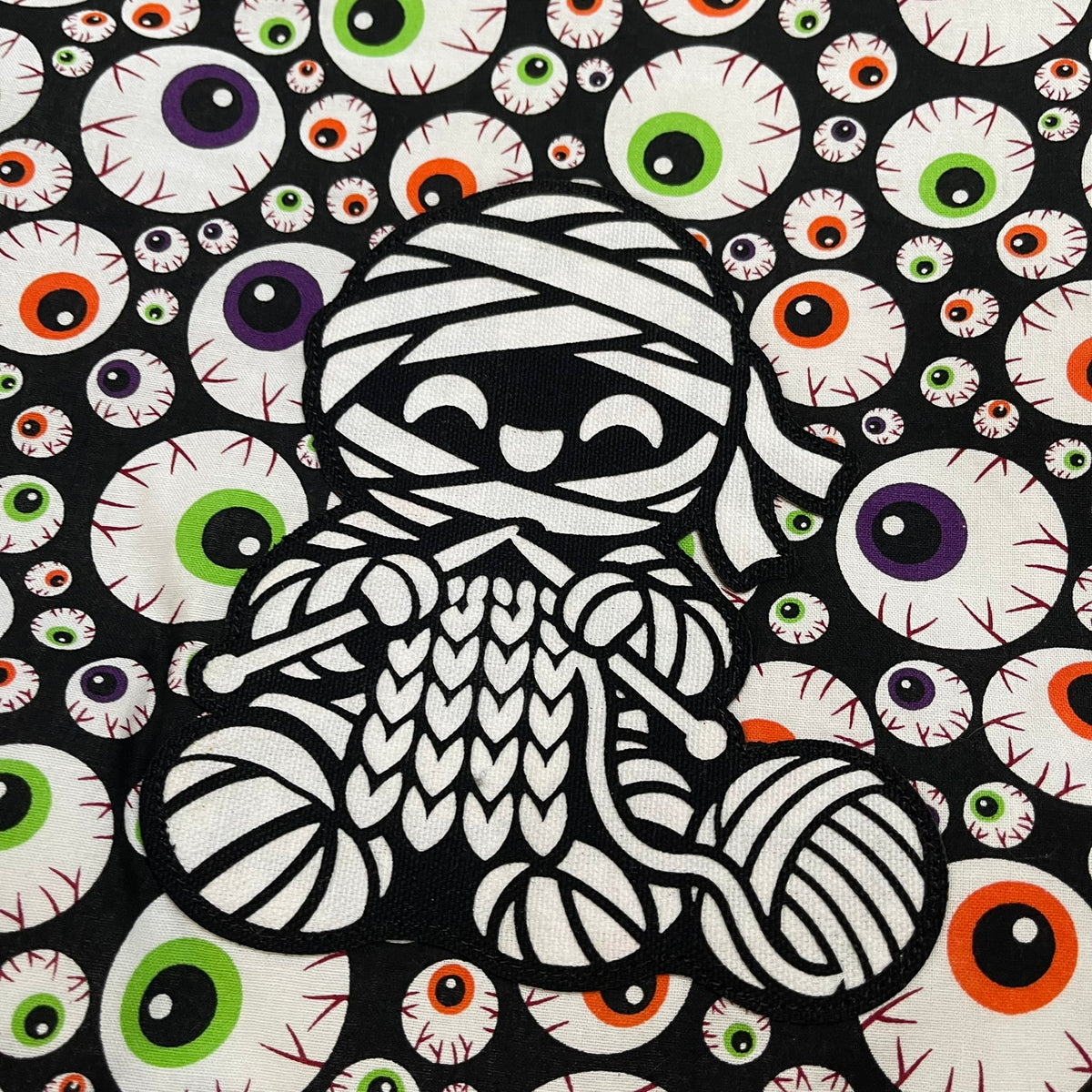 Close-up of a smiling mummy appliqué knitting beside a ball of yarn on colorful eyeball-print fabric.
